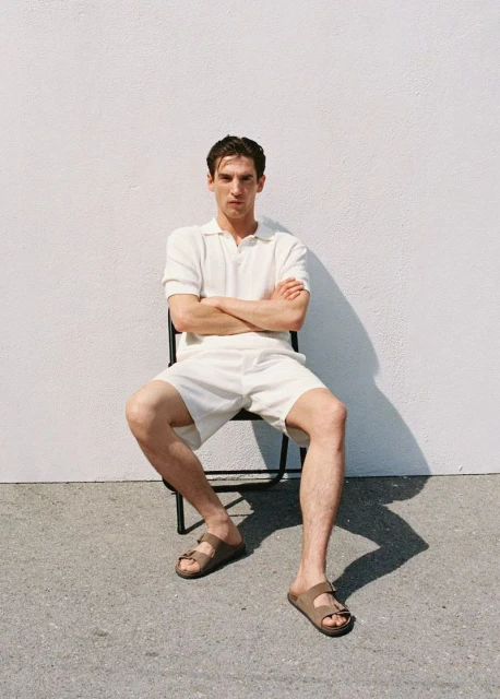 Adult male in off-white polo and shorts sitting on a chair outside, wearing sandals, with arms crossed against a white wall.