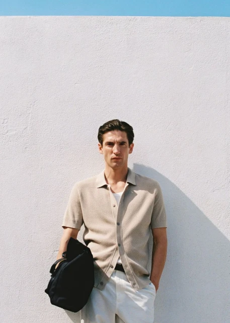 An adult male stands against a bright white textured wall wearing a beige button-down shirt, white pants, and holding a black backpack.