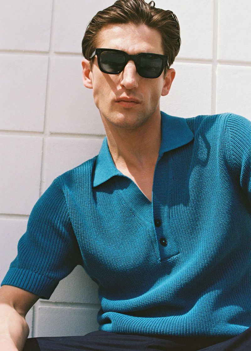 Man in sunglasses and teal polo shirt poses against tiled wall. Outdoor setting with casual fashion and cool, modern style.
