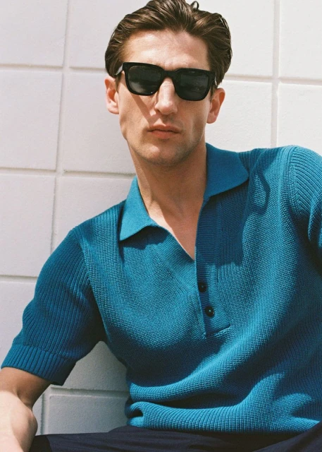 Man in sunglasses and teal polo shirt poses against tiled wall. Outdoor setting with casual fashion and cool, modern style.