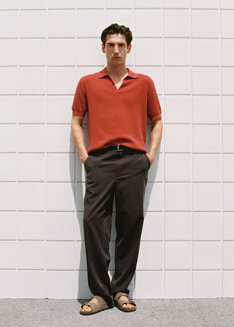 Man in red polo shirt and dark trousers, wearing sandals, standing against a grid-patterned wall in an eye-level full shot.