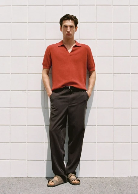 Man in red polo shirt and dark trousers, wearing sandals, standing against a grid-patterned wall in an eye-level full shot.