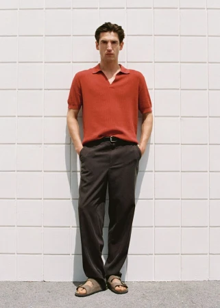 Man in red polo shirt and dark trousers, wearing sandals, standing against a grid-patterned wall in an eye-level full shot.