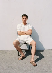 Model seated on a black chair wearing a white polo shirt and white shorts against a textured white wall.