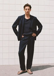 A man wearing a dark linen suit and sandals posing against a white tiled wall for Mango