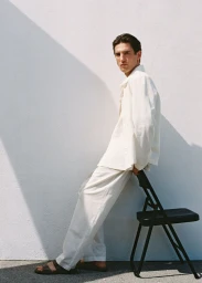 Model leaning against a black chair wearing white linen shirt and trousers against a white wall.