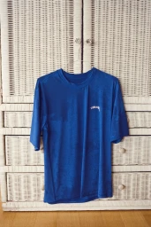 Royal blue Stussy short-sleeve t-shirt hanging on a wire hanger in front of a white woven wicker wardrobe.