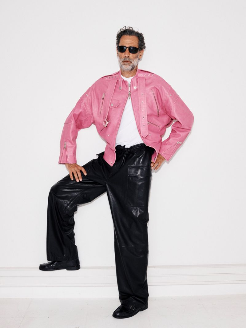 Model John Armstrong wearing a pink leather moto jacket, black wide-leg trousers, and sunglasses against a white wall.