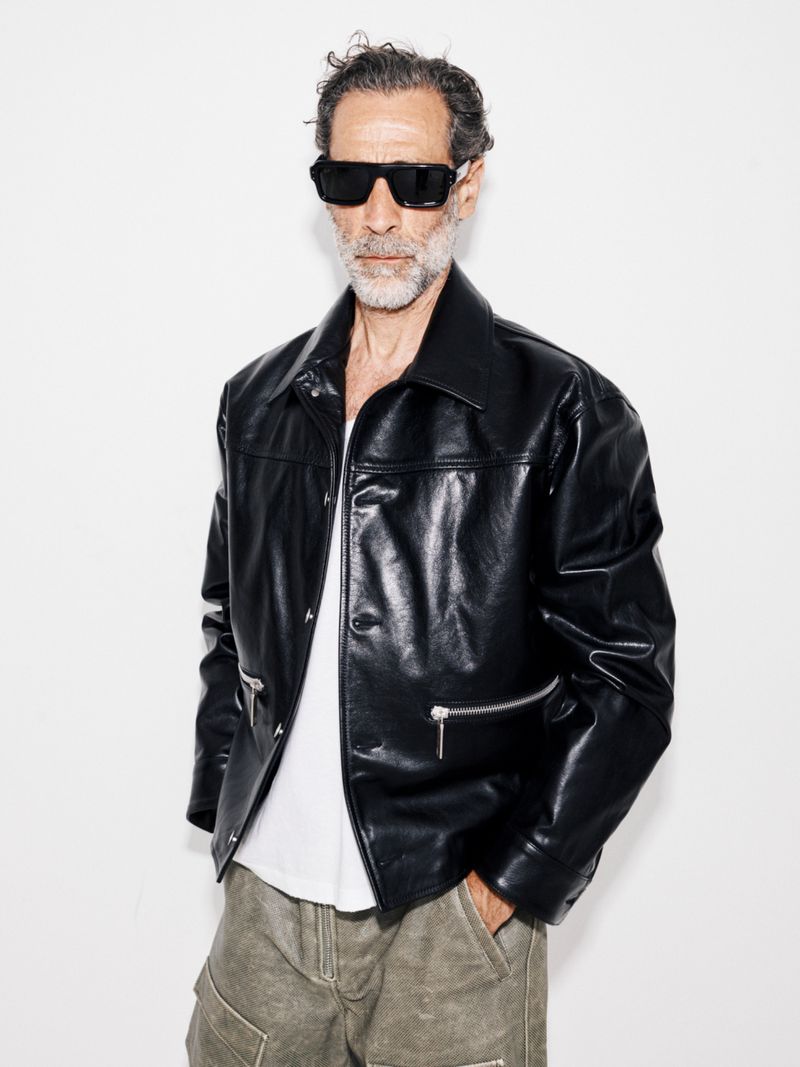 John Armstrong wearing black sunglasses and a shiny black leather jacket against a white studio background.