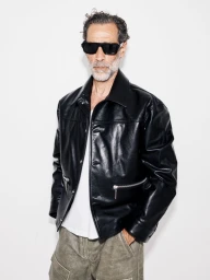 John Armstrong wearing black sunglasses and a shiny black leather jacket against a white studio background.