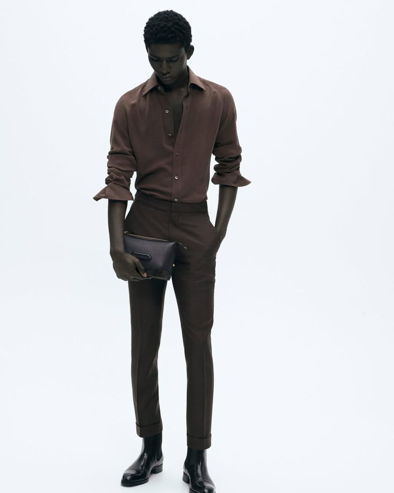 Dark-skinned male fashion model wearing brown, long-sleeved shirt, brown trousers, and black Chelsea boots, holding dark handbag against white backdrop.