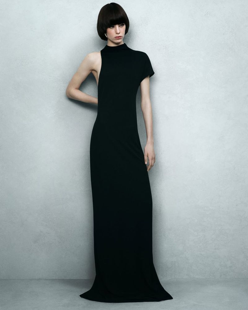 A slender woman posing in a long, asymmetrical black dress with one sleeve against a textured gray wall, showcasing simple elegance.