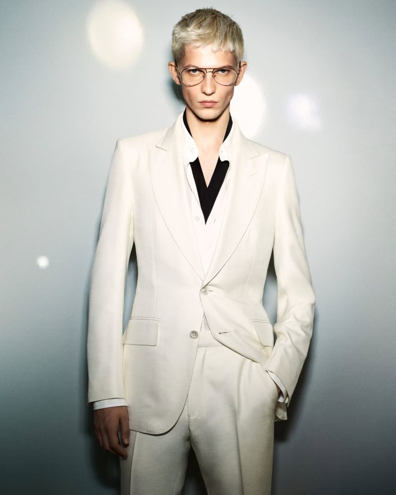 Man with blond hair poses in a white suit and aviator glasses against a neutral background with bokeh light flares.