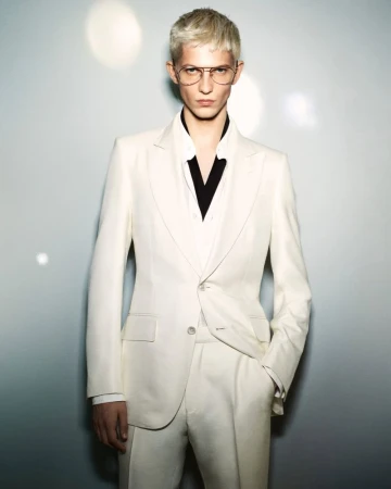 Man with blond hair poses in a white suit and aviator glasses against a neutral background with bokeh light flares.