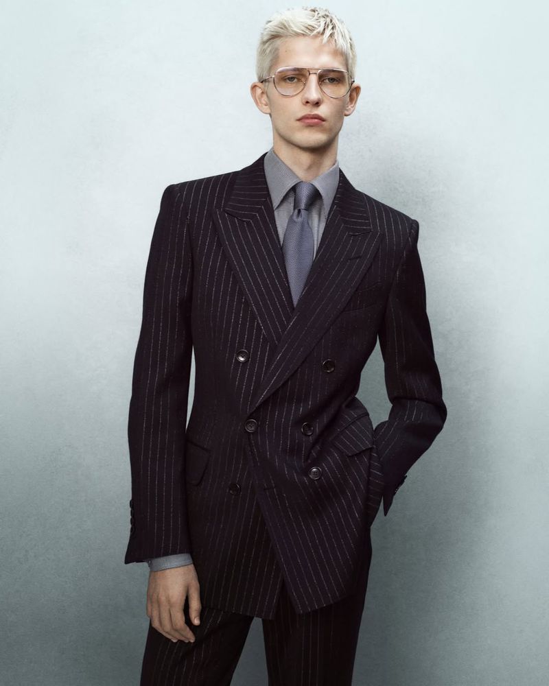Portrait of a young adult male with blond hair wearing clear glasses, gray dress shirt and tie, and a striped black double-breasted suit.
