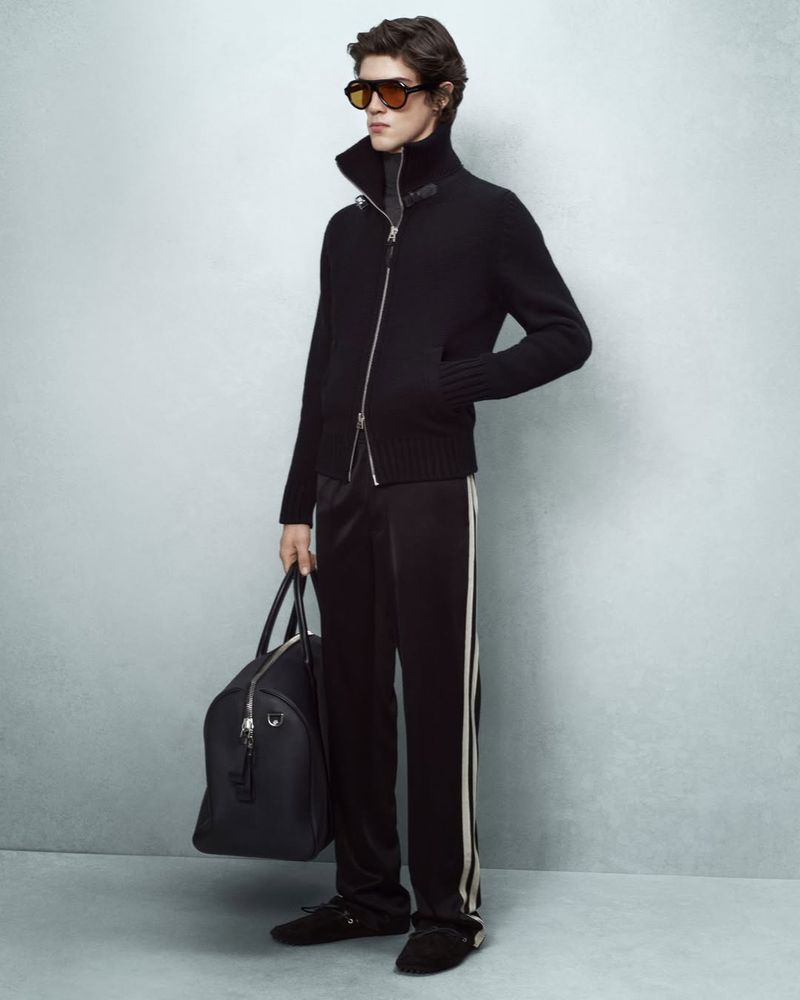 Male model in black zip-up jacket and sunglasses holding a black duffel bag against a grey seamless backdrop