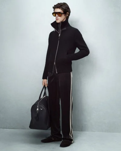 Male model in black zip-up jacket and sunglasses holding a black duffel bag against a grey seamless backdrop