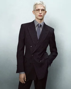 Model with bleached hair wearing a dark pinstripe double-breasted suit against a grey studio backdrop