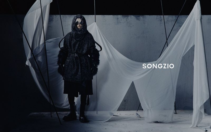 Model wearing an oversized hooded black textured coat standing amid draped white translucent fabric on a studio set.