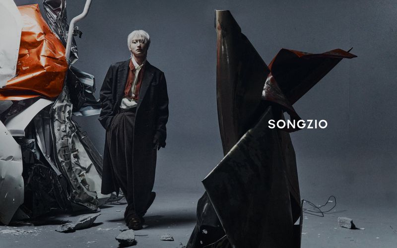 Park Seonghwa standing in a dark coat and trousers against a grey studio backdrop featuring crushed metal debris.