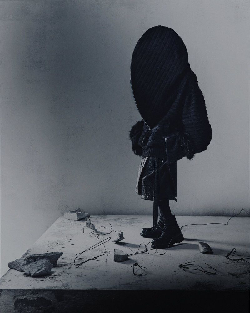 Model wearing a sculptural black hooded outfit and boots on a concrete platform with wire debris for Songzio.