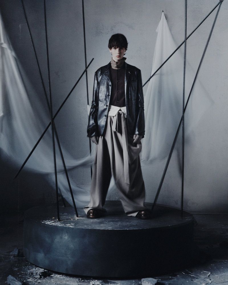Model wearing a dark leather jacket and draped wide-leg trousers on a round plinth for the Songzio campaign.