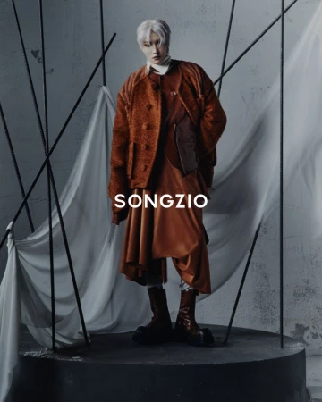 Park Seonghwa wearing a textured rust jacket and draped skirt trousers standing on a round platform for Songzio.