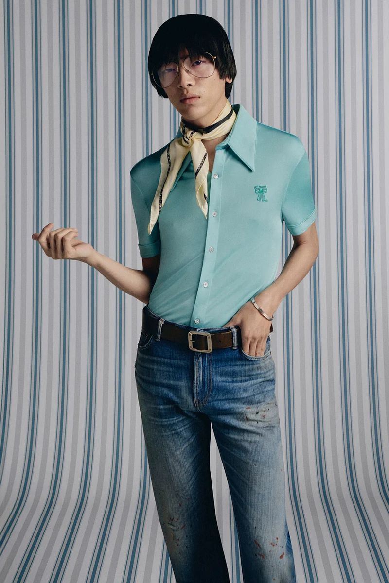 Studio shot of a slender adult male with glasses and a scarf, wearing a turquoise collared shirt and distressed jeans against a striped backdrop.
