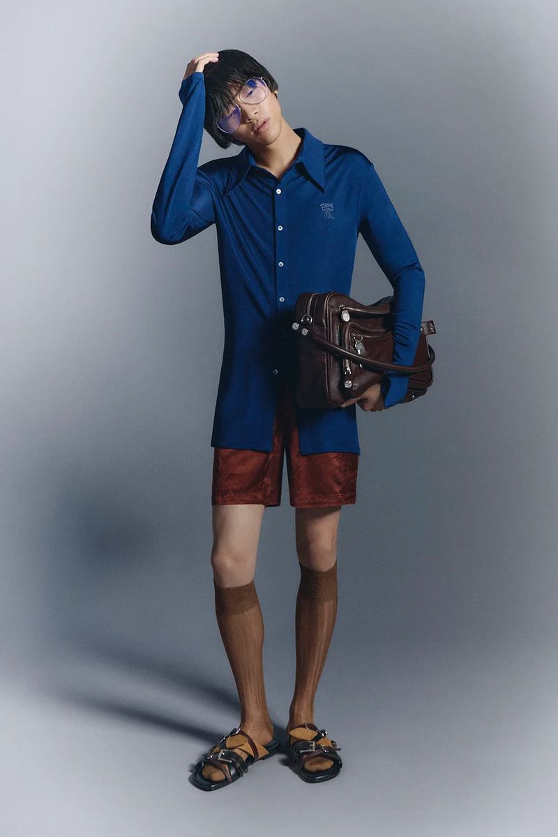 A slender man in a blue shirt, red shorts, brown knee socks, and sandals poses with a leather bag against a grey studio backdrop.
