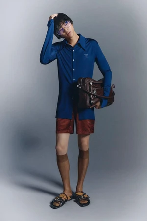 Model wearing blue long-sleeve shirt, red shorts, and brown bag against a grey background