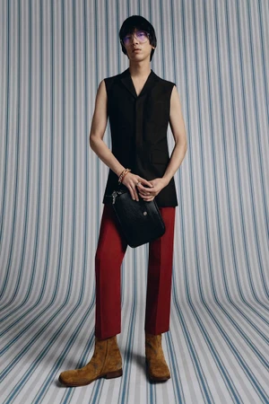Model wearing a black sleeveless vest and red trousers holding a black pouch against a striped backdrop