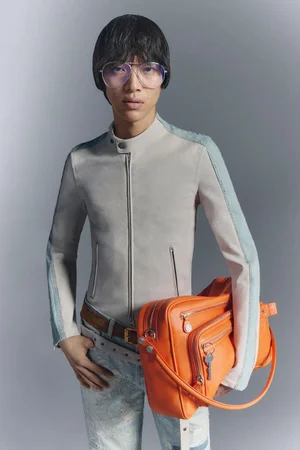 Model Akito Mizutani wearing a cream jacket and tinted glasses holding an orange bag against a grey seamless background