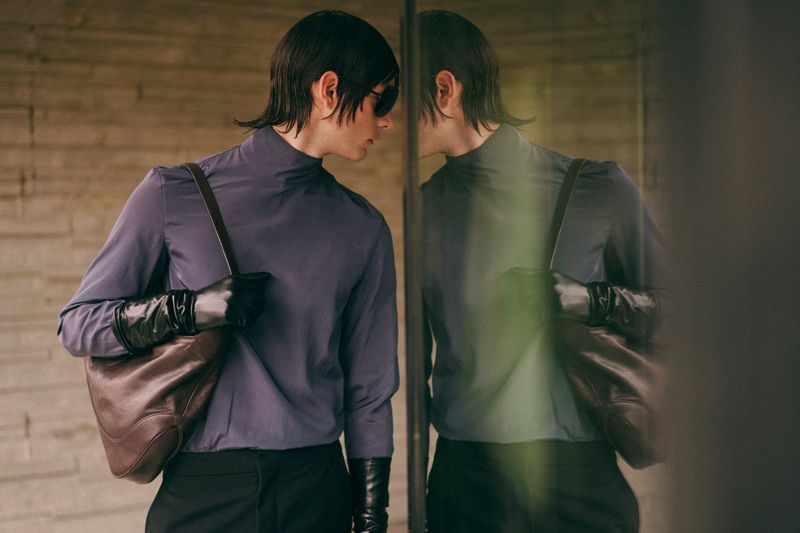 Model wearing a slate blue turtleneck, black leather gloves, and dark sunglasses carrying a brown leather bag next to a glass reflection.