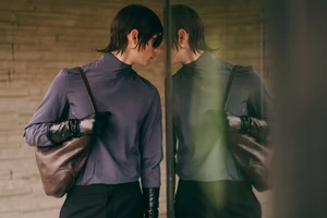 Model wearing a slate blue turtleneck, black leather gloves, and dark sunglasses carrying a brown leather bag next to a glass reflection.