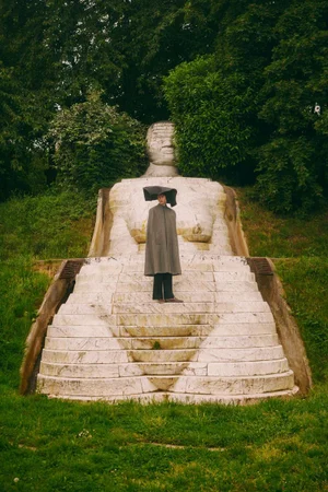 Model wearing a grey cape and sculptural black headpiece standing on a monumental stone statue surrounded by green park trees.