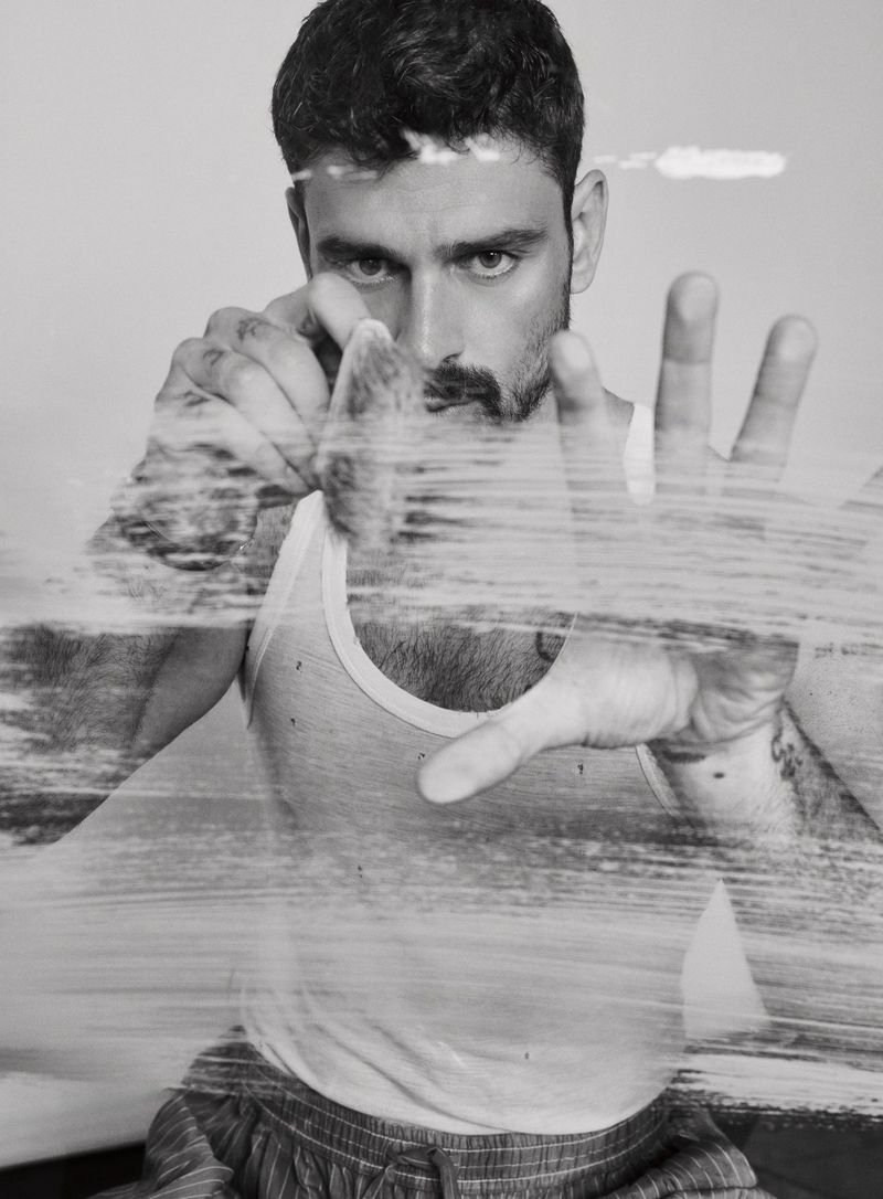 Close-up of Michele Morrone in a white tank top with hand pressed against a glass surface with smeared texture, captured in black and white.
