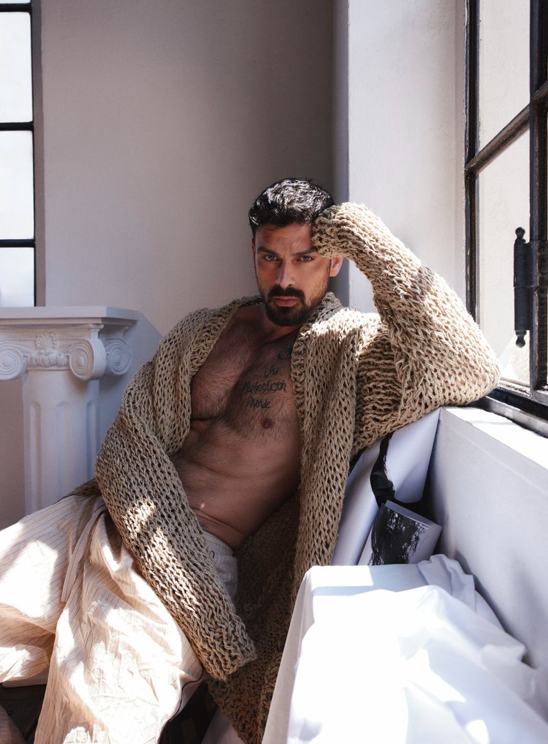 Michele Morrone wearing a beige chunky knit cardigan open over a bare chest, seated indoors beside a window with bright natural light.