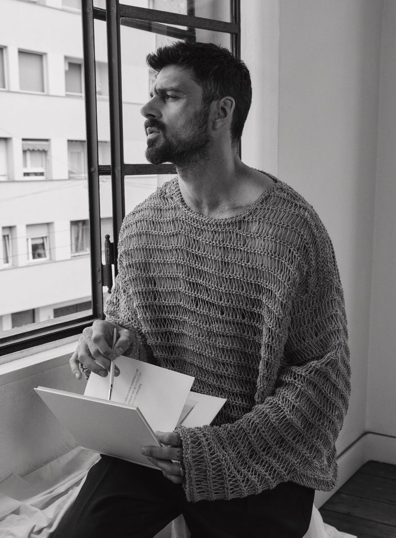 Man with beard wearing an open-knit oversized grey sweater looking out of a window while holding a small sketchbook and a pen.