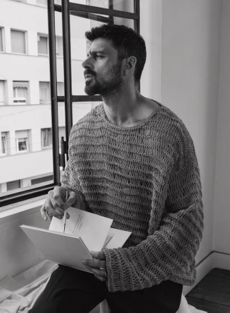 Man with beard wearing an open-knit oversized grey sweater looking out of a window while holding a small sketchbook and a pen.