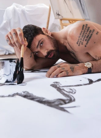 Shirtless man with tattoos lying on the floor holding a paintbrush over a white canvas with black ink strokes in an art studio.