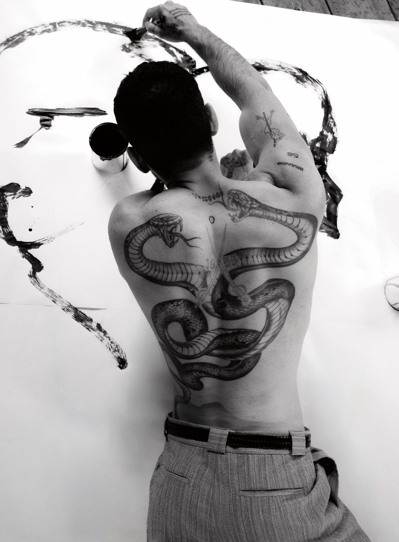Shirtless man with a large snake tattoo on his back painting with black ink on a large white sheet of paper