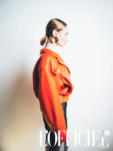 Profile view of a woman wearing a voluminous bright orange leather jacket and a pleated gold earring against a white backdrop.