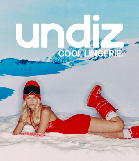 Model lying on artificial snow in a red bodysuit, red beanie, ski goggles, and red moon boots in front of a snowy mountain backdrop.