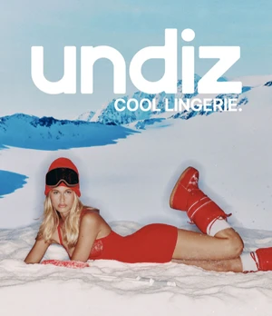Model lying on artificial snow in a red bodysuit, red beanie, ski goggles, and red moon boots in front of a snowy mountain backdrop.
