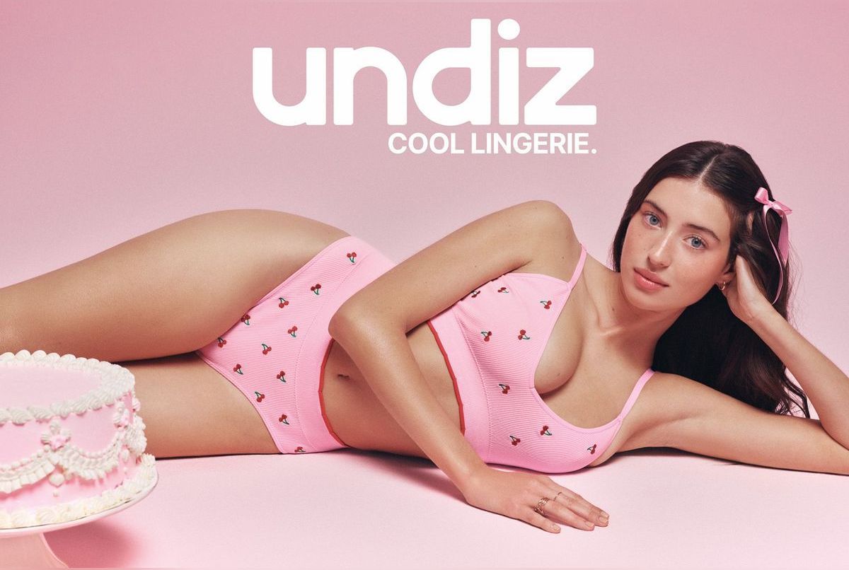 Model reclining in a pink cherry-patterned lingerie set beside a decorated cake against a pink studio backdrop.