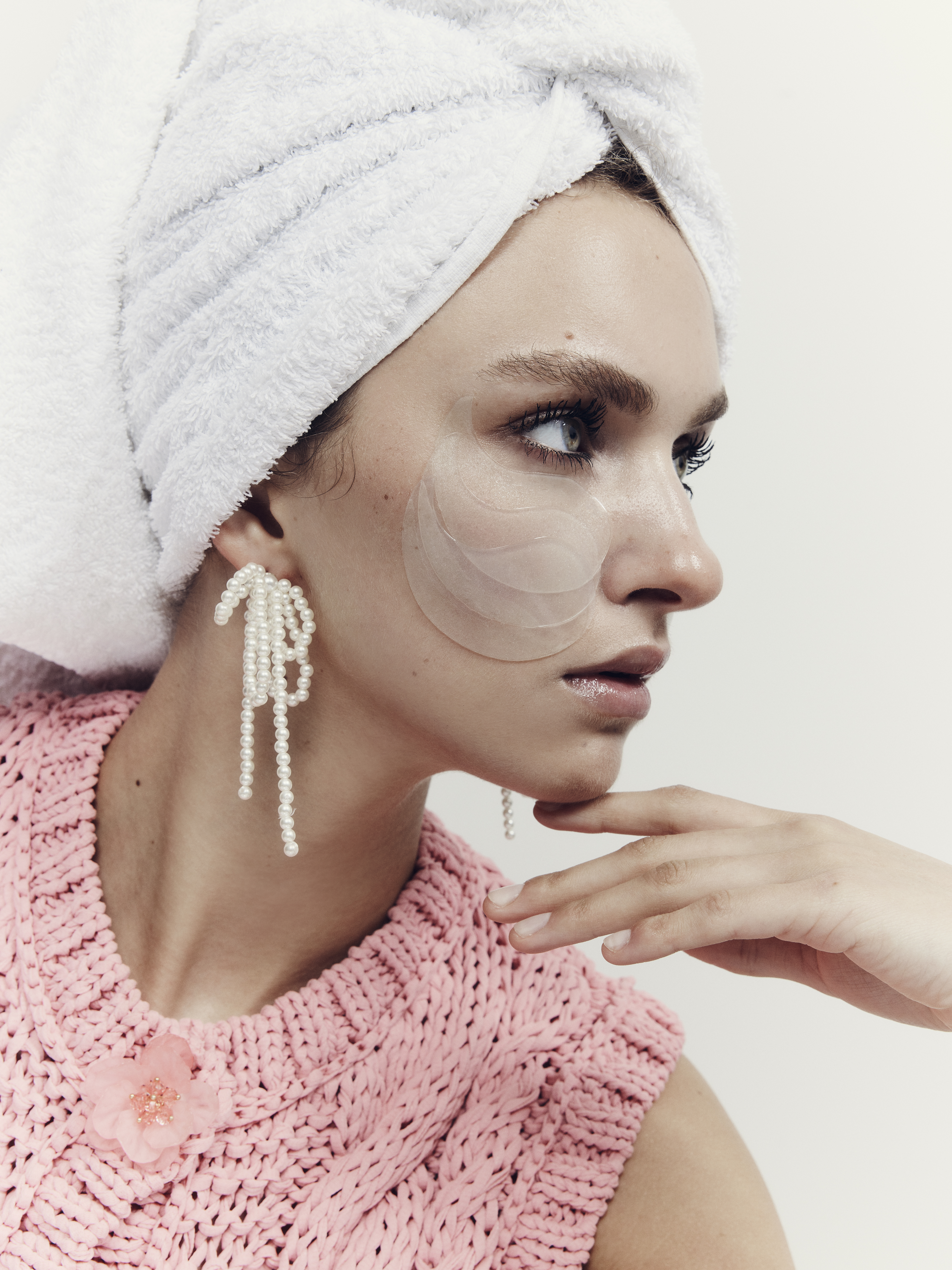 A woman with a white towel wrapped around her head wearing pearl bow earrings and a pink knit top with eye patches on her face.