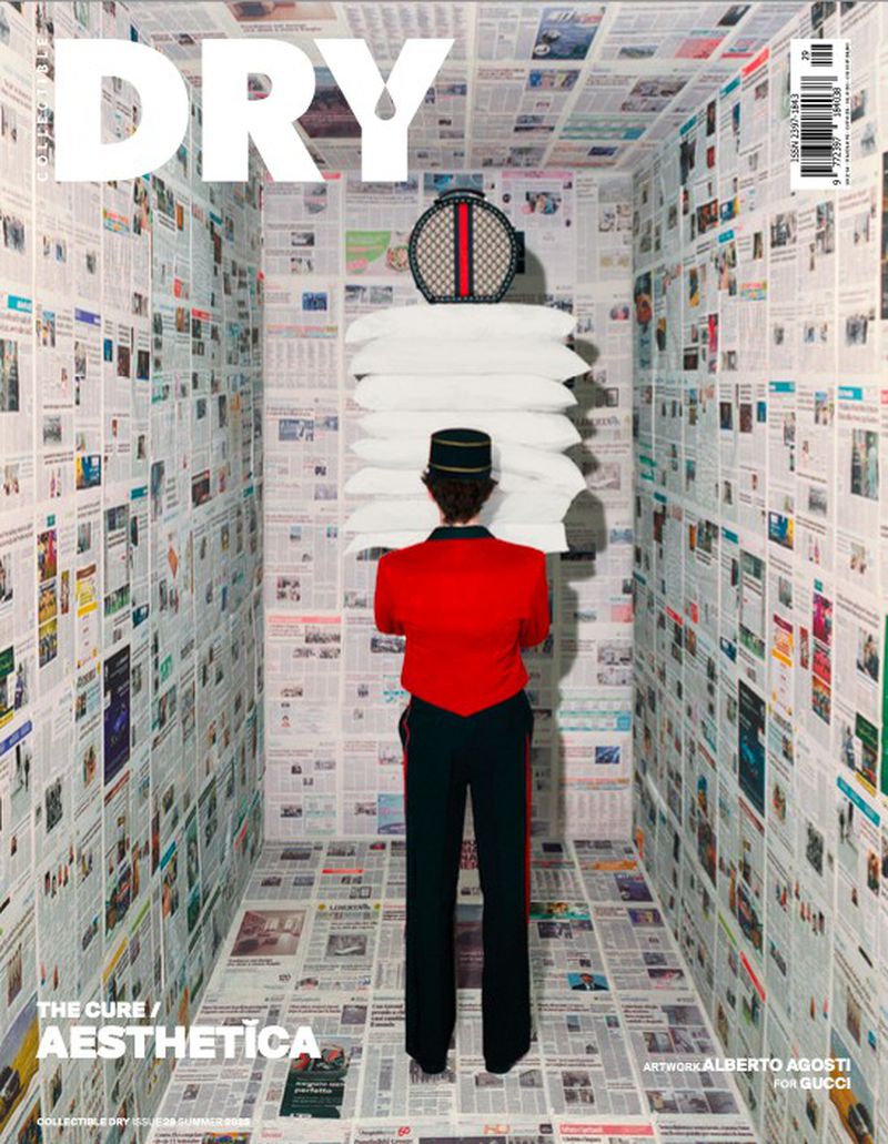 Magazine cover features a person in a hotel uniform carrying pillows and a bag in a newspaper-covered room, showcasing a surreal editorial style.
