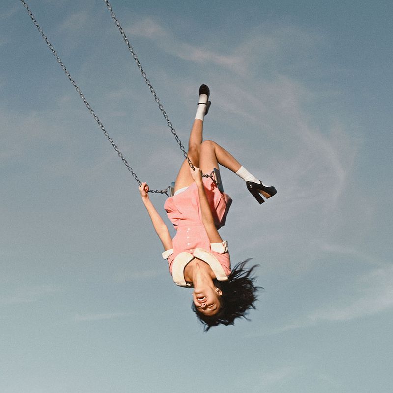 A woman hanging upside down on a swing against a clear blue sky wearing a pale pink dress, white socks, and black platform Mary Jane shoes.