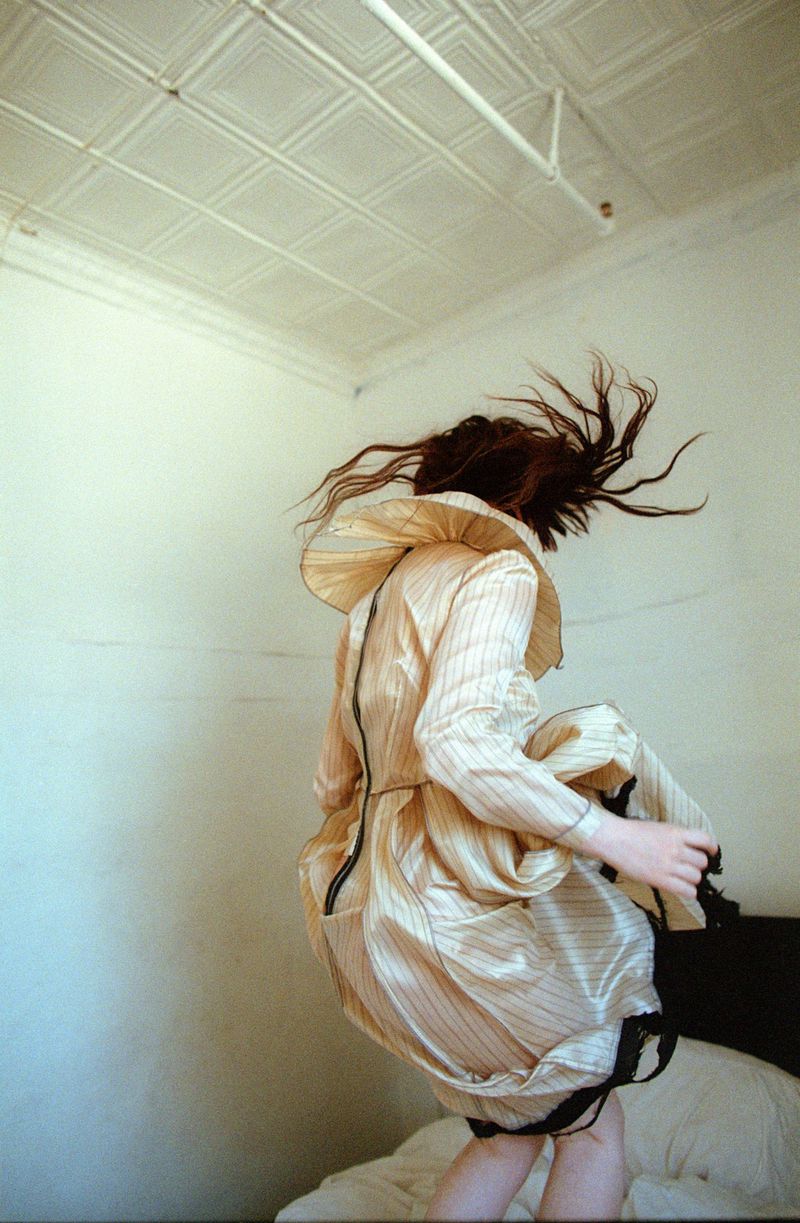 Sophie Thatcher jumping on a bed in a pleated beige striped dress with hair tossing upward in an editorial portrait.