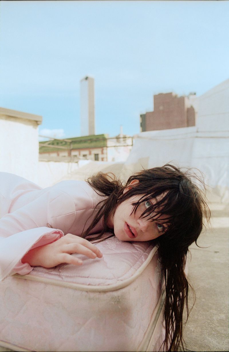 Sophie Thatcher lying on a pink mattress on an outdoor rooftop wearing a light pink coat.